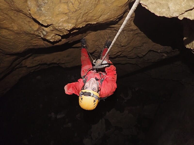 Spéléologue en combinaison rouge et casque jaune descend en rappel dans une cavité sombre et rocheuse.