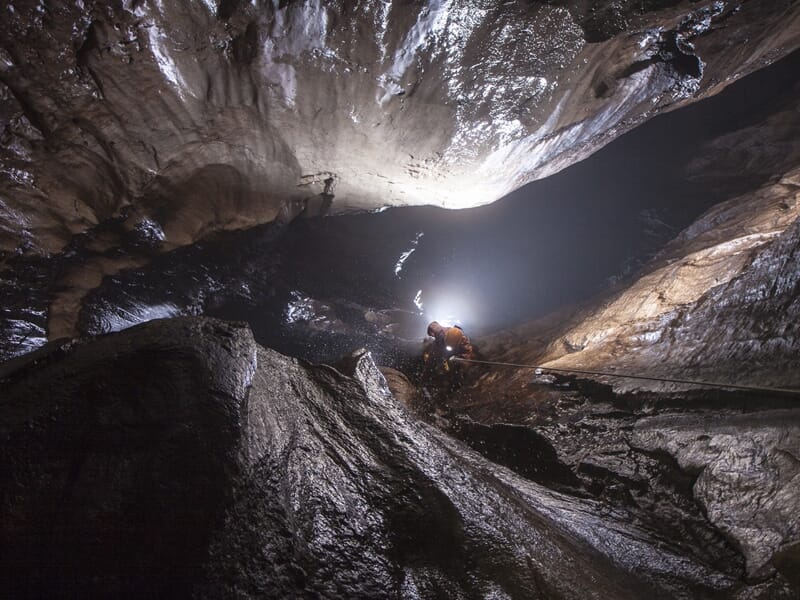 Spéléologue explore une vaste grotte sombre éclairée par une lampe frontale, entouré de parois rocheuses humides.