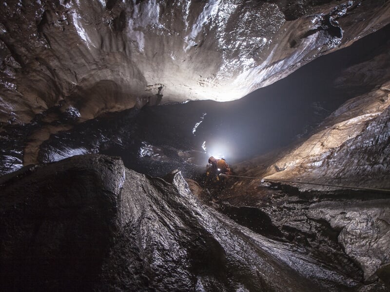 Spéléologue explore une vaste grotte sombre éclairée par une lampe frontale, entouré de parois rocheuses humides.