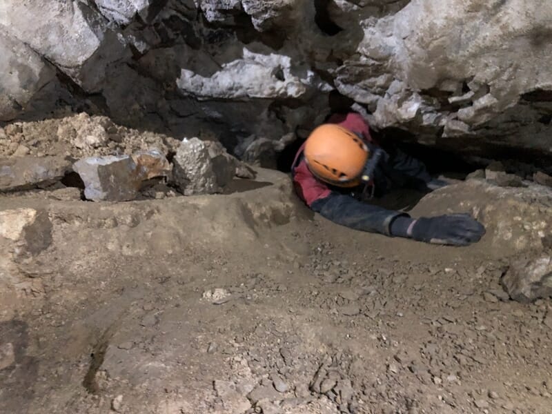 Spéléologue équipé d'un casque orange progressant dans une étroite galerie rocheuse souterraine.