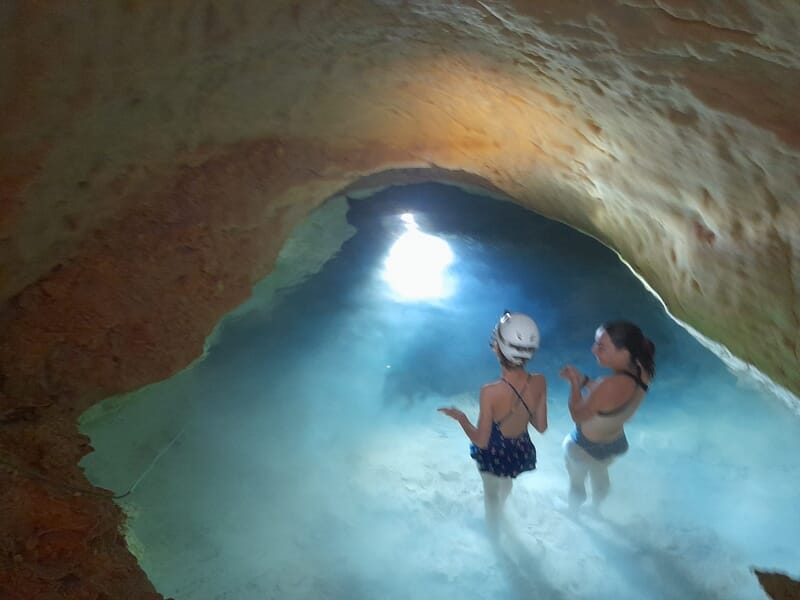 Deux enfants en maillot et casque blanc dans une grotte, observant une source lumineuse dans l'eau.
