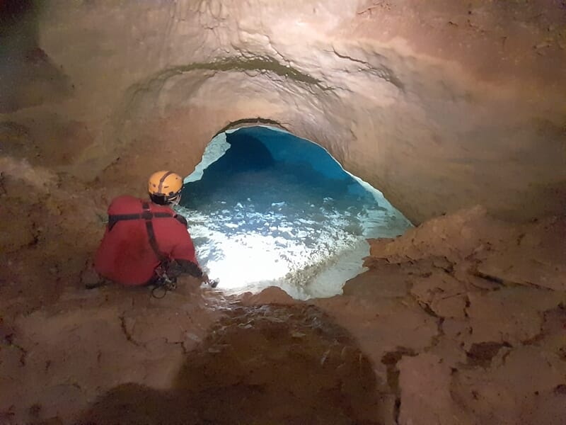Spéléologue en combinaison rouge assis à l'entrée d'une cavité éclairée par une lumière bleue.