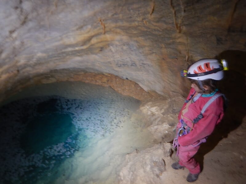 Spéléologue en combinaison rose et casque blanc regardant une vasque d'eau claire dans une grotte.