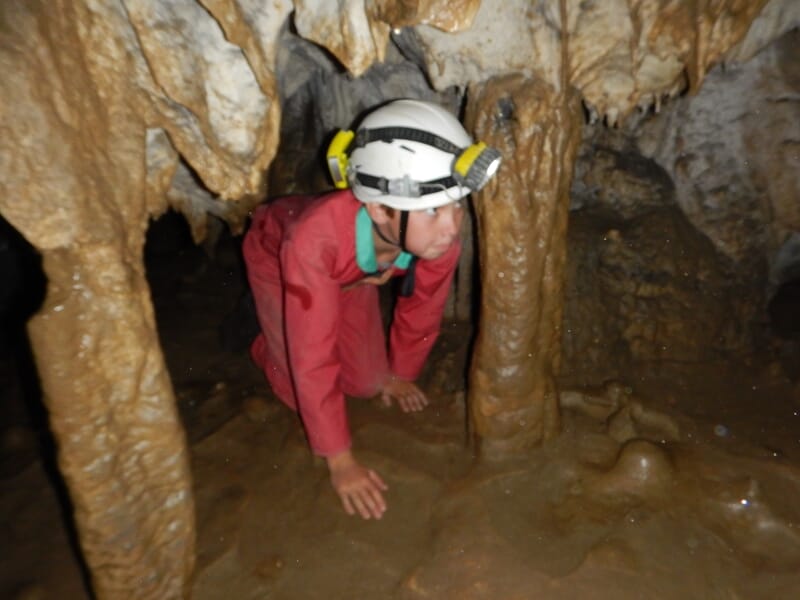 Spéléologue en combinaison rouge et casque avec lampe explore une grotte étroite entre des formations rocheuses humides.