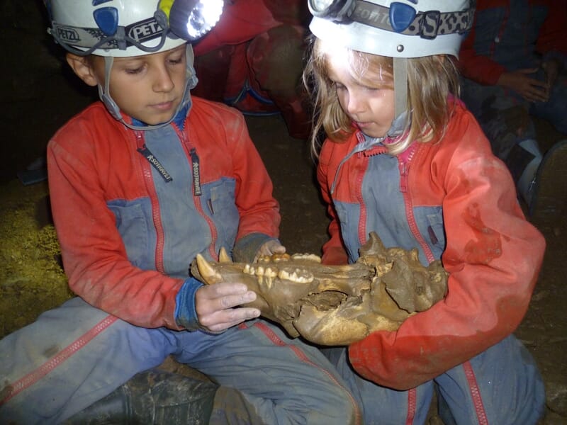 Deux enfants en combinaison rouge et casque examinent un grand os fossilisé dans une grotte souterraine.