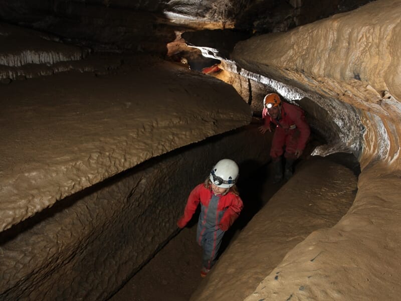 Deux spéléologues en combinaison rouge et casque marchent dans un passage étroit et sinueux d'une grotte.