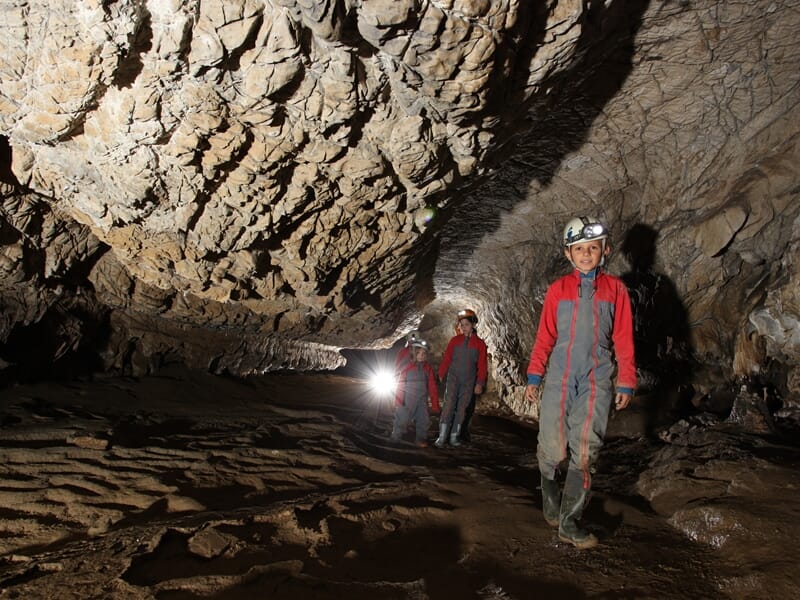 Groupe de spéléologues en combinaison rouge explore une grande salle rocheuse avec des lampes frontales allumées.