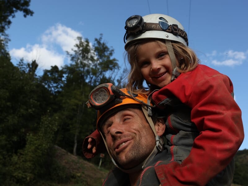 Un homme portant un casque avec lampe porte une petite fille souriante en combinaison rouge sur ses épaules en extérieur.