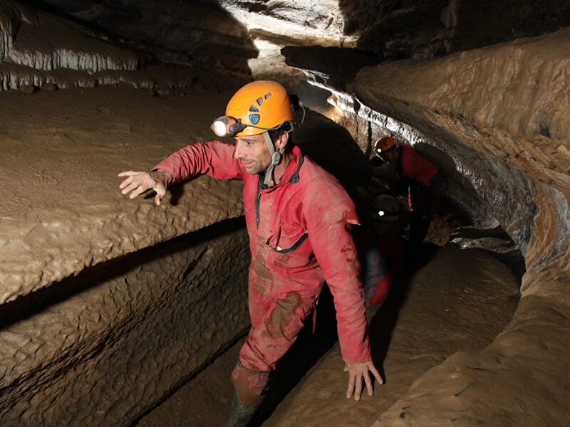 Spéléologue en combinaison rouge et casque avec lampe avance prudemment dans un passage étroit d'une grotte rocheuse.
