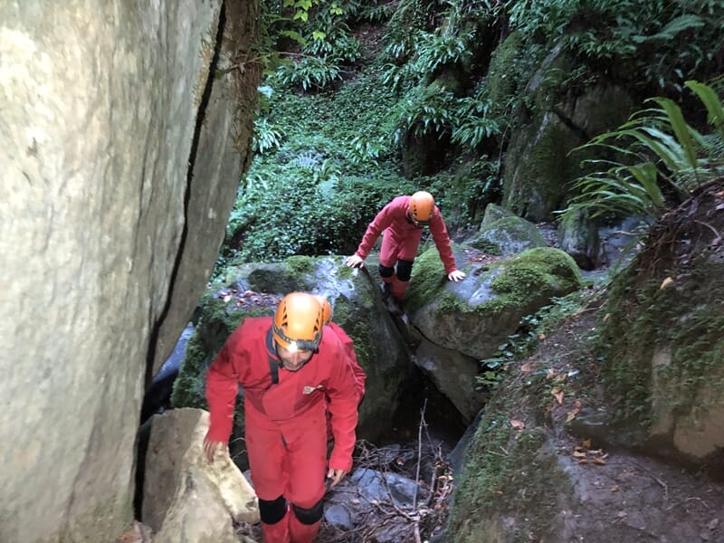 Deux spéléologues en combinaison rouge et casque jaune grimpent entre des rochers dans une forêt dense et humide.
