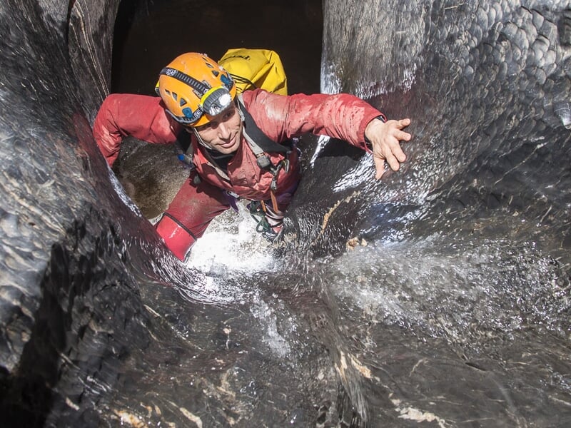 Spéléologue en combinaison rouge et casque jaune descend une paroi rocheuse lisse et humide dans une grotte.