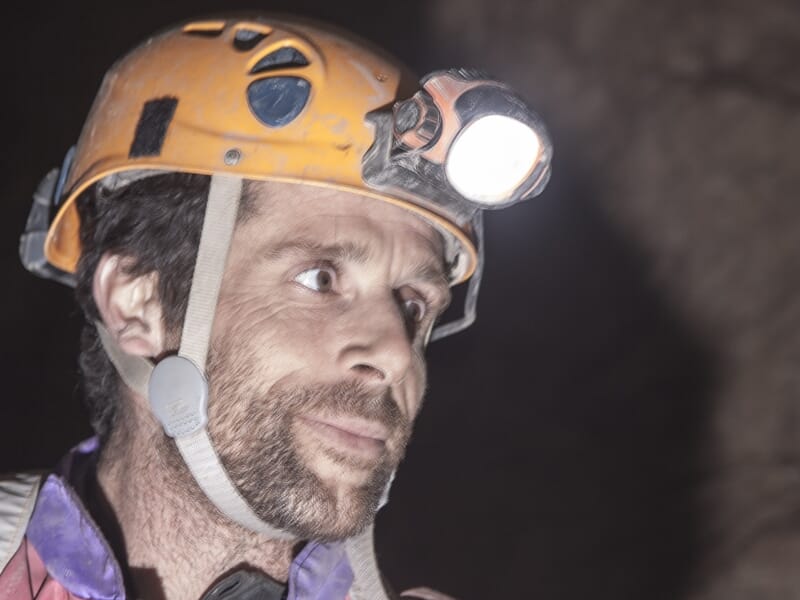 Portrait d'un spéléologue avec casque jaune et lampe frontale, regardant vers la droite dans une grotte sombre.