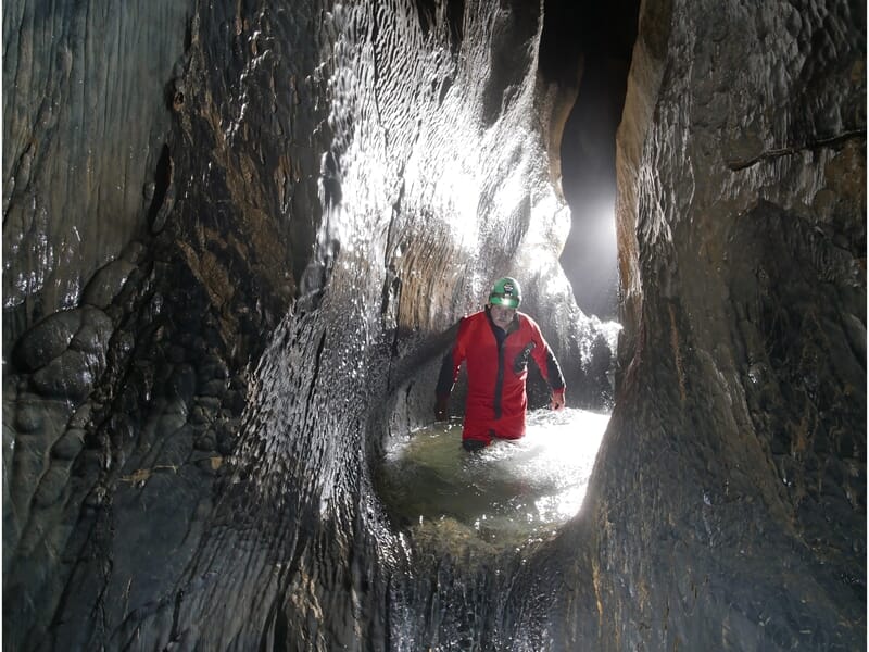 Spéléologue en combinaison rouge et casque vert marche dans une rivière souterraine étroite aux parois lisses.