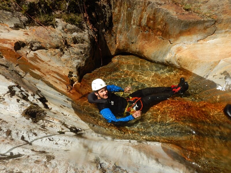 Homme souriant en combinaison de canyoning glisse sur une roche lisse dans un bassin d'eau transparente.