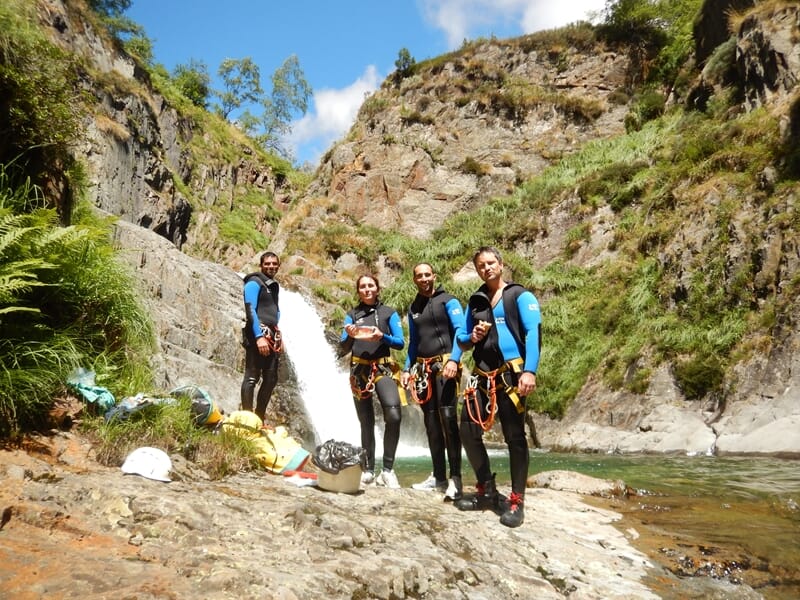 Groupe de quatre personnes en tenue de canyoning posent devant une cascade dans un paysage montagneux.