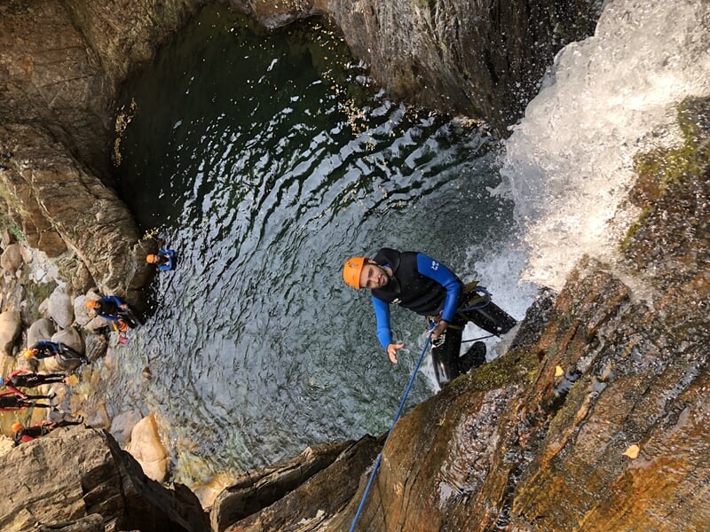 Personne en casque orange descend en rappel le long d'une cascade dans un canyon rocheux avec un bassin.