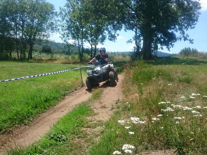 Personne en quad roulant sur un chemin de terre entouré d'herbes et de fleurs blanches sous un ciel bleu clair.