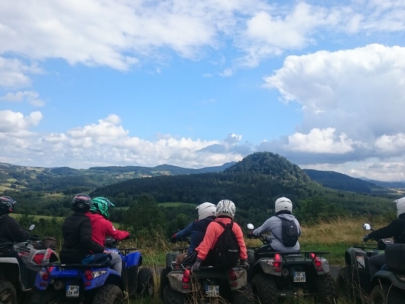 Groupe de personnes en quad regardant un paysage vallonné avec collines et ciel nuageux en arrière-plan.