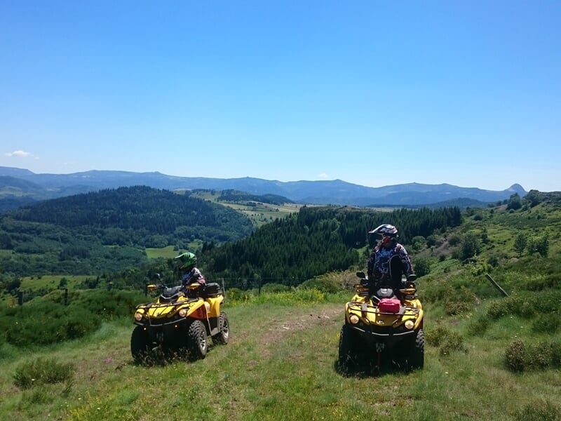 Deux personnes en quad jaunes sur une colline avec une vue dégagée sur une vallée verdoyante et un ciel bleu.