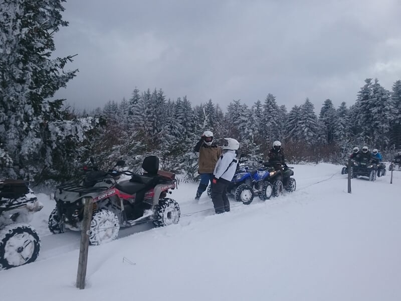 Groupe de quads et personnes en tenue hivernale sur un chemin enneigé bordé d'arbres sous un ciel gris.