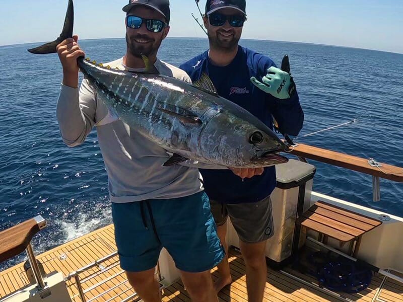 Deux hommes souriants sur un bateau tenant un gros thon pêché, mer calme en arrière-plan.