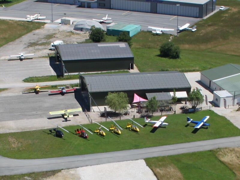 Vue aérienne d'un aérodrome avec plusieurs petits avions légers alignés sur l'herbe devant des hangars.