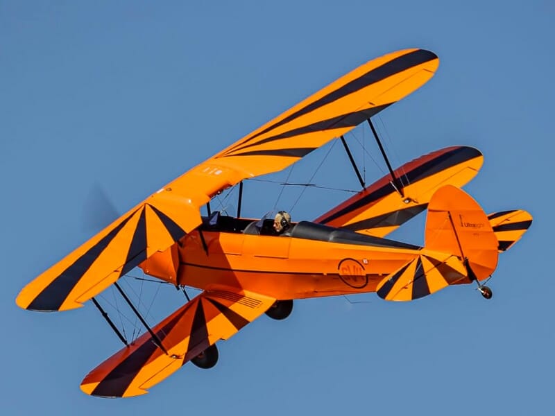 Avion biplan orange avec motifs noirs en vol sous un ciel bleu clair, pilote visible dans le cockpit ouvert.