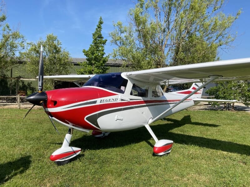 Petit avion Legend 600 et blanc stationné sur une pelouse verte avec des arbres en arrière-plan.