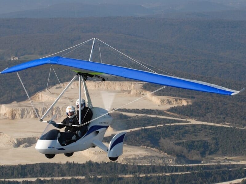 ULM pendulaire biplace blanc avec une aile bleue en vol au-dessus d'une zone de carrières lors d'un baptême de l'air en ULM près de Dijon.