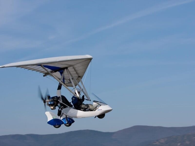 Un ULM pendulaire biplace blanc et bleu en vol au-dessus d'un paysage montagneux sous un ciel clair et dégagé.