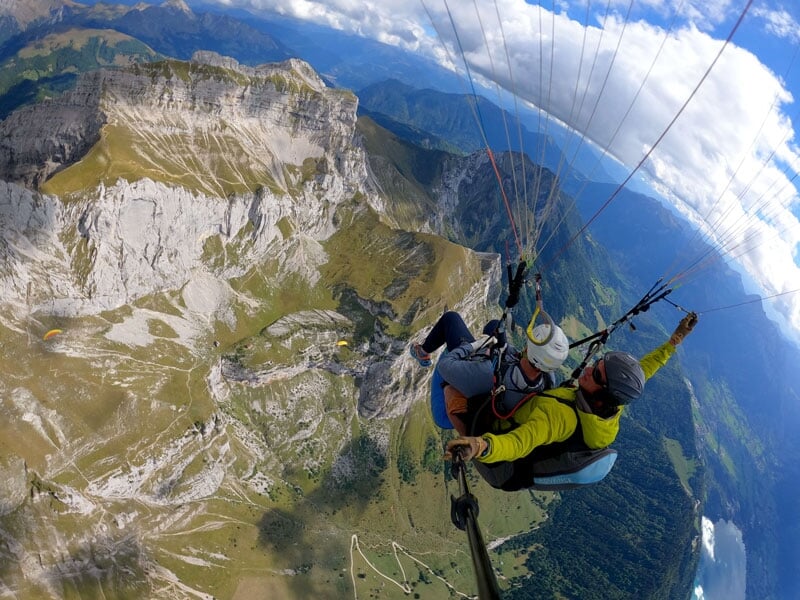 Vue impressionnante d'un parapente en tandem naviguant au-dessus de paysages montagneux escarpés, dominés par des formations rocheuses blanches et vertes. Les parapentistes, vêtus de jaune et bleu, se