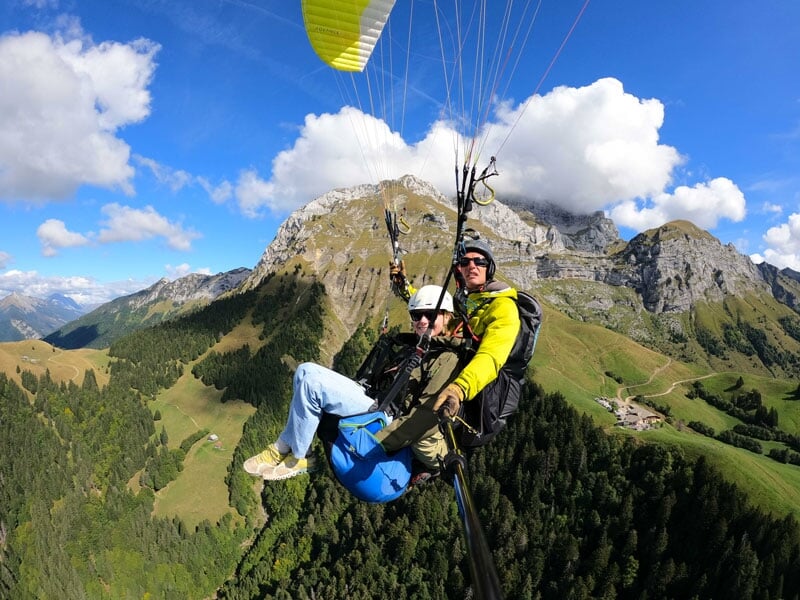 Parapente en tandem au-dessus de vallées verdoyantes et de montagnes, avec des nuages bas touchant les sommets. L'instructeur et son passager, vêtus de jaune et bleu, profitent d'une vue dégagée sur u