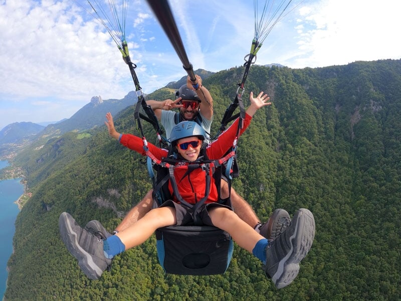 Scène dynamique de parapente au-dessus d'un paysage de lac et de forêts, avec un passager en rouge exprimant l'excitation par un geste de joie. Le cadre montagneux et la vaste étendue d'eau en contreb
