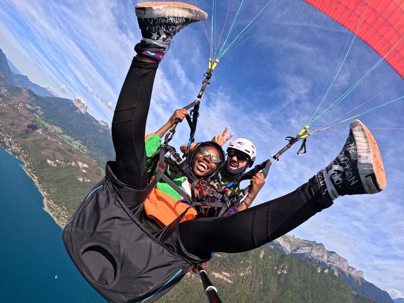 Image captivante de deux parapentistes exprimant un enthousiasme débordant, avec l'un des passagers réalisant une figure acrobatique. Le paysage panoramique comprend un grand lac et des montagnes loin