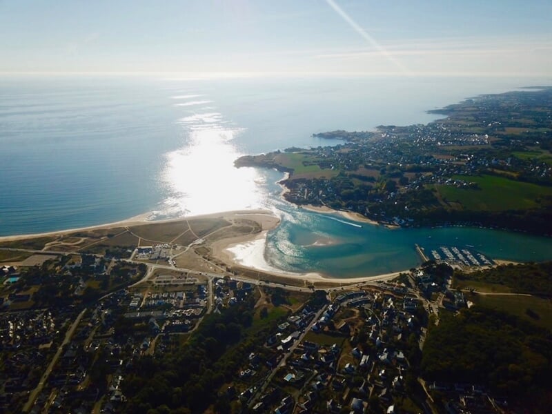Vue aérienne de la plage de Guidel avec l'océan, le soleil se reflétant sur l'eau et la côte verdoyante en arrière-plan.