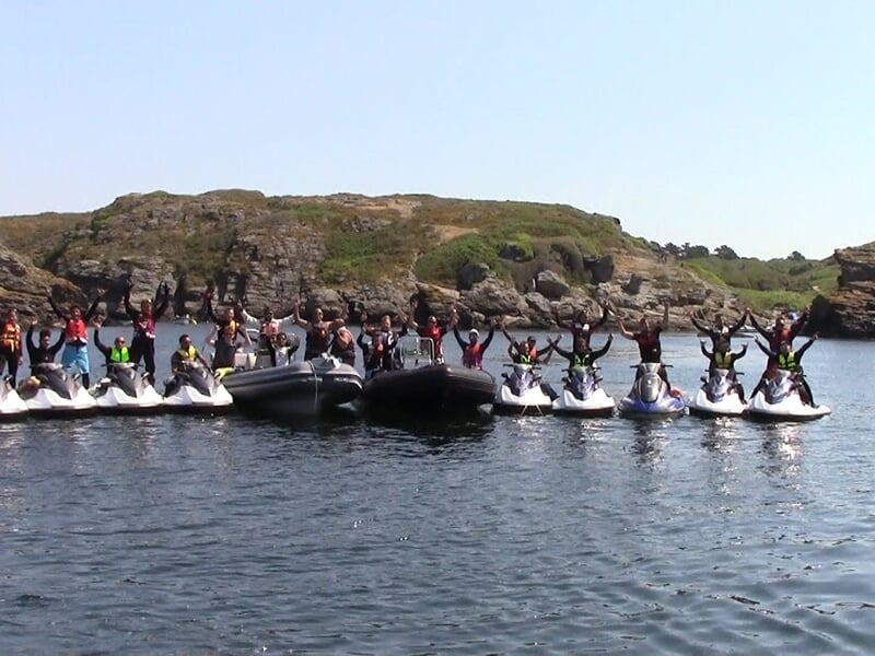 Groupe de personnes sur des jetskis alignés sur l'eau, levant les bras en signe de joie près d'une côte rocheuse.