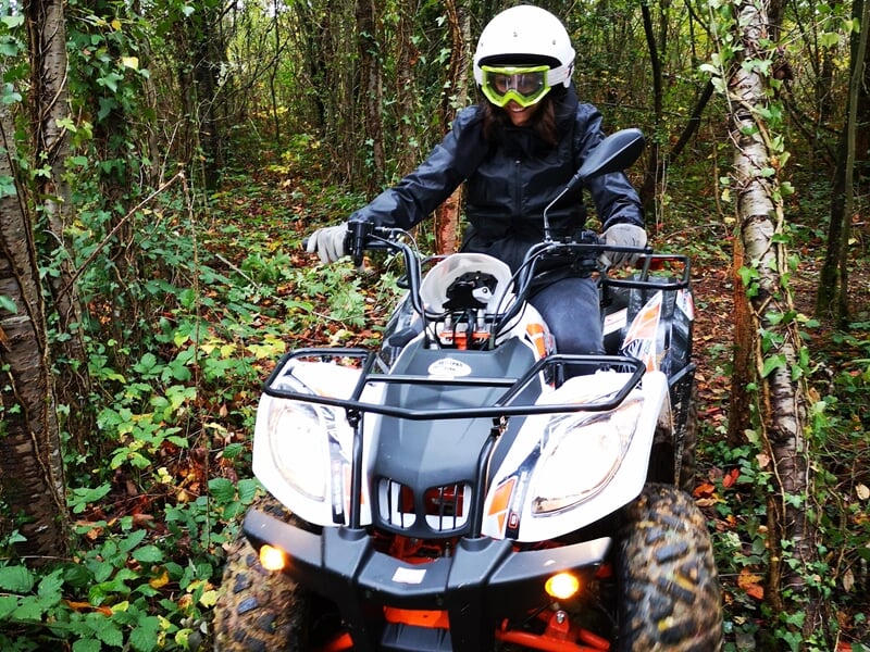 Personne en tenue de protection et casque blanc conduisant un quad orange dans un sentier forestier dense.