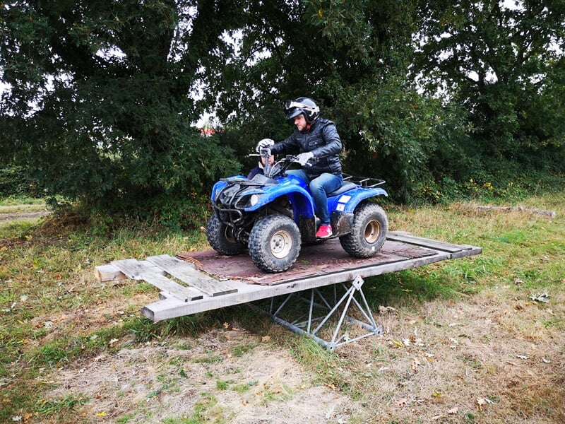 Conducteur sur un quad bleu franchissant un obstacle en bois sur un terrain herbeux avec un grand arbre.