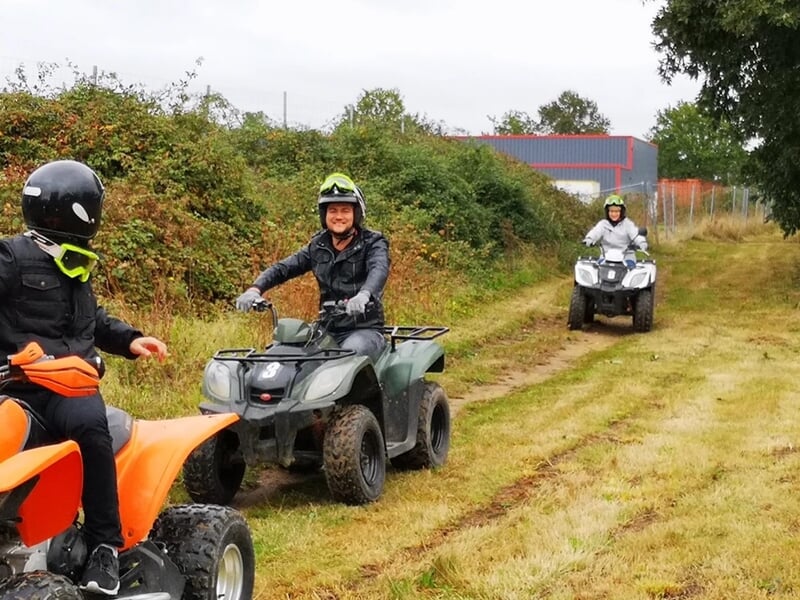 Groupe de personnes en quads roulant sur un chemin herbeux avec des buissons et un bâtiment en arrière-plan.