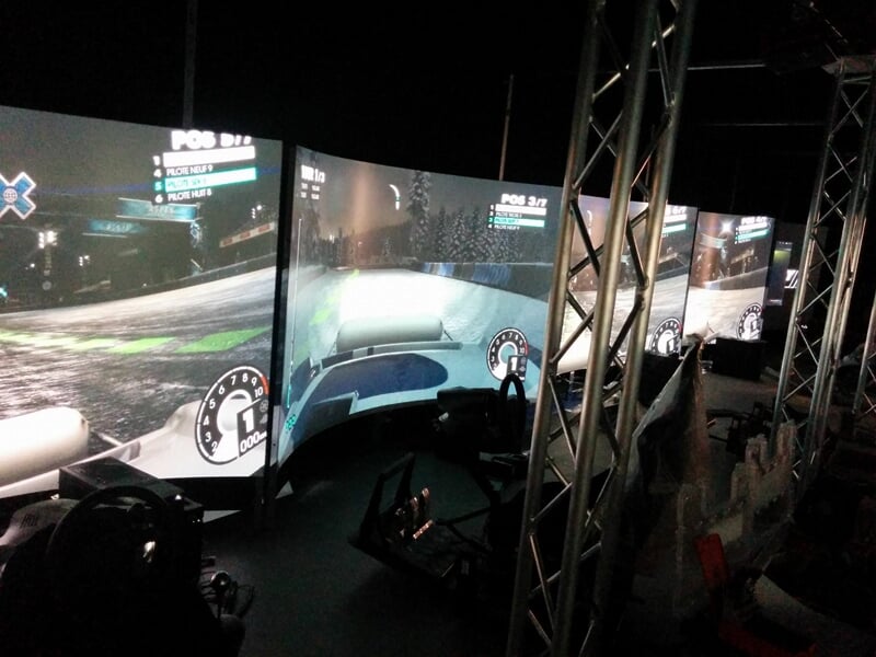 Plusieurs simulateurs de conduite automobile alignés avec écrans incurvés et volants dans une salle sombre.