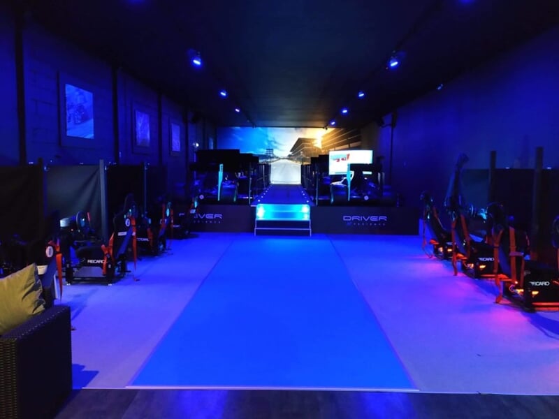 Salle sombre avec plusieurs simulateurs de conduite alignés, éclairage bleu et ambiance futuriste.