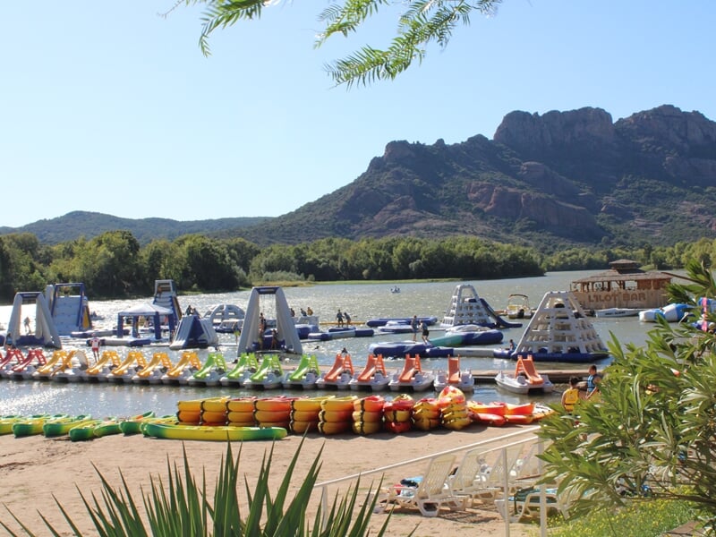 Plage avec rangées de pédalos colorés et structures gonflables sur l’eau, montagnes verdoyantes sous ciel clair.