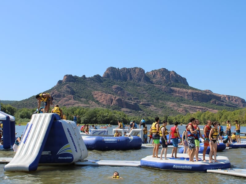 Structures gonflables aquatiques avec enfants en gilets jaunes, toboggan, montagnes rocheuses en arrière-plan et ciel bleu dégagé.