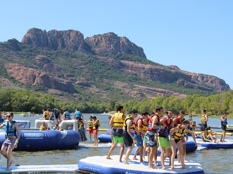 Groupe de jeunes en gilets sur plateformes gonflables flottantes, montagnes rocheuses en fond et eau calme.