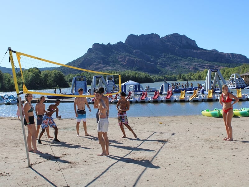 Groupe de jeunes en maillots et shorts jouant au volley sur plage de sable, structures gonflables et montagnes en fond.