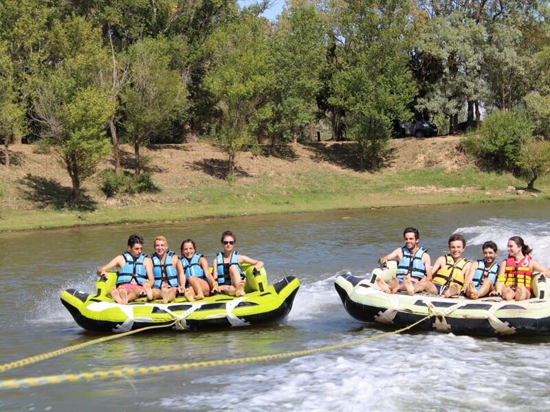 Deux bouées tractées côte à côte transportant des groupes de jeunes en gilets colorés sur une rivière bordée d’arbres.