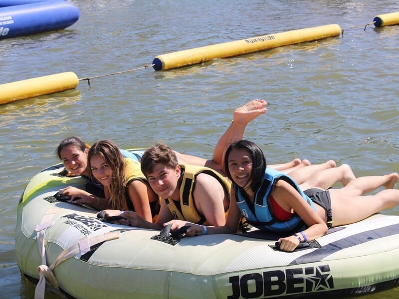 Quatre jeunes allongés sur une bouée flottante près d’un parc aquatique, souriants et en gilets de sauvetage colorés.