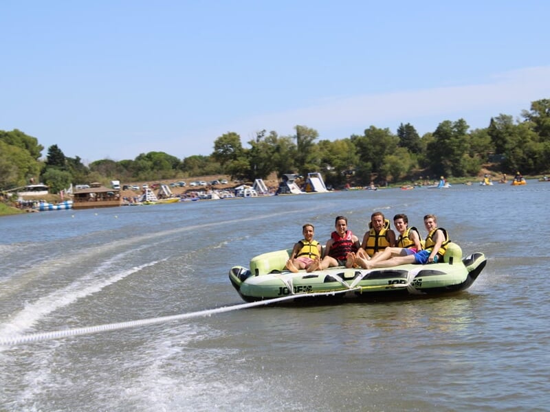 Groupe de six personnes en gilets de sauvetage sur une bouée tractée glissant sur un plan d’eau bordé de structures gonflables.