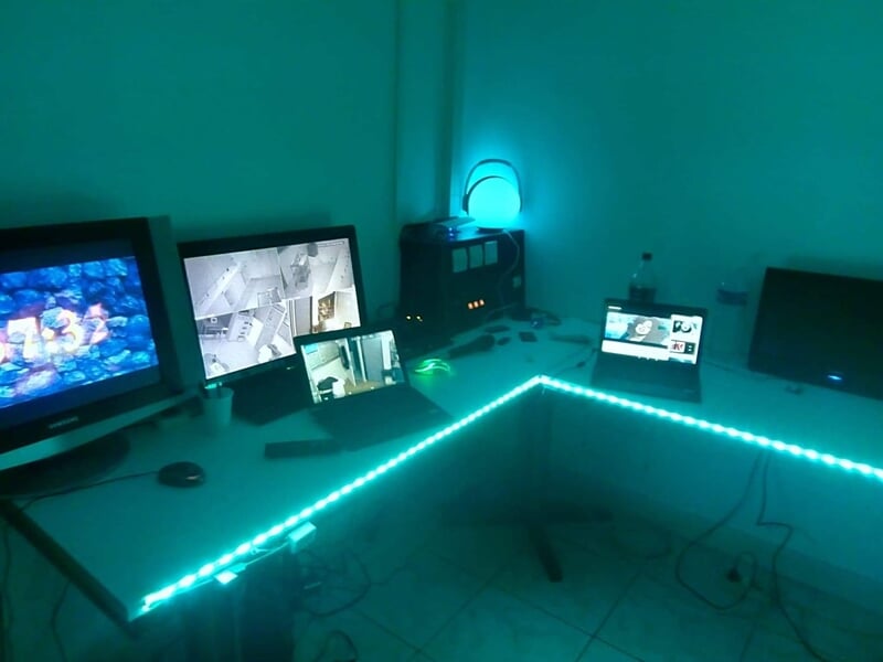 Bureau en angle avec plusieurs écrans et ordinateurs, éclairage bleu et bande LED sous le bureau dans une salle de contrôle.