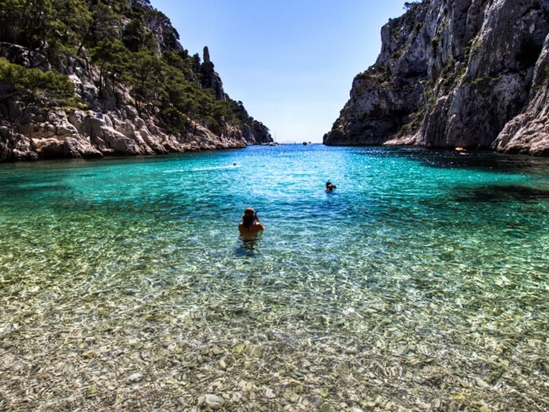 Personnes nageant dans une crique aux eaux turquoise entourée de falaises rocheuses sous un ciel bleu clair.